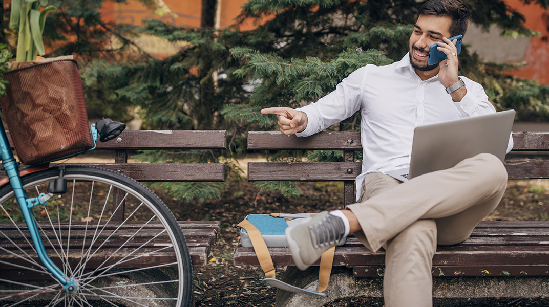 Young mail professional working remotely outdoors with laptop and phone, reflecting flexibility needed for summer corporate housing and business travel.